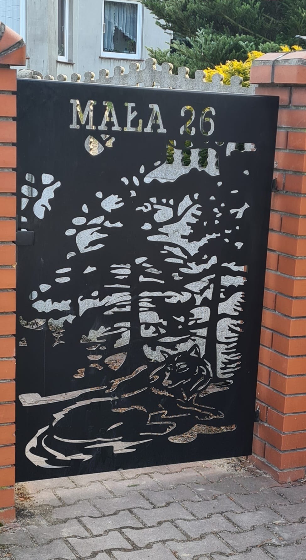 Czarna, metalowa brama z wycinanym laserowo motywem wilka w lesie i adresem 'MAŁA 26', osadzona w ceglanym murze.