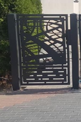 Nowoczesna, metalowa furtka z ażurowym wzorem, wykonana w Kamyku, z widocznymi spawami i elementami dekoracyjnymi przypominającymi gałęzie drzew.