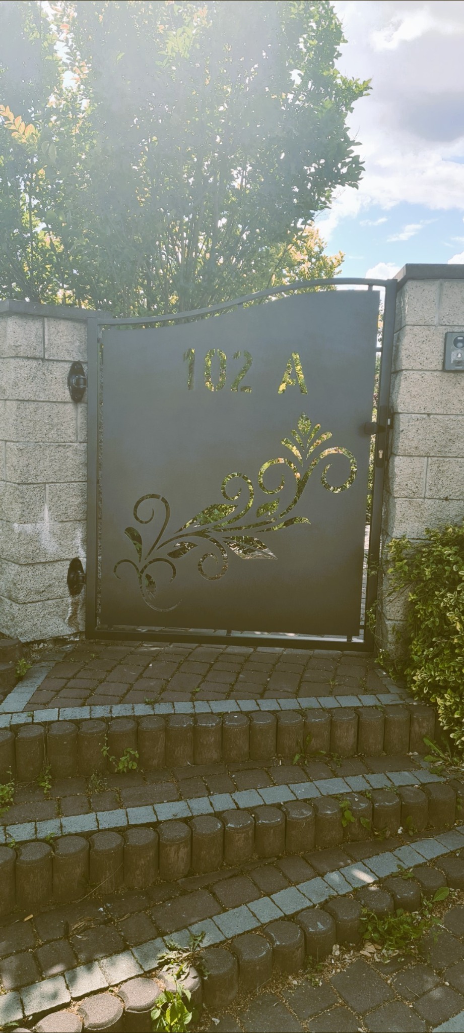 Metalowa brama z wycinanym laserowo wzorem roślinnym i numerem domu 102 A, zamontowana na murowanych słupkach, prowadząca do schodów wejściowych.