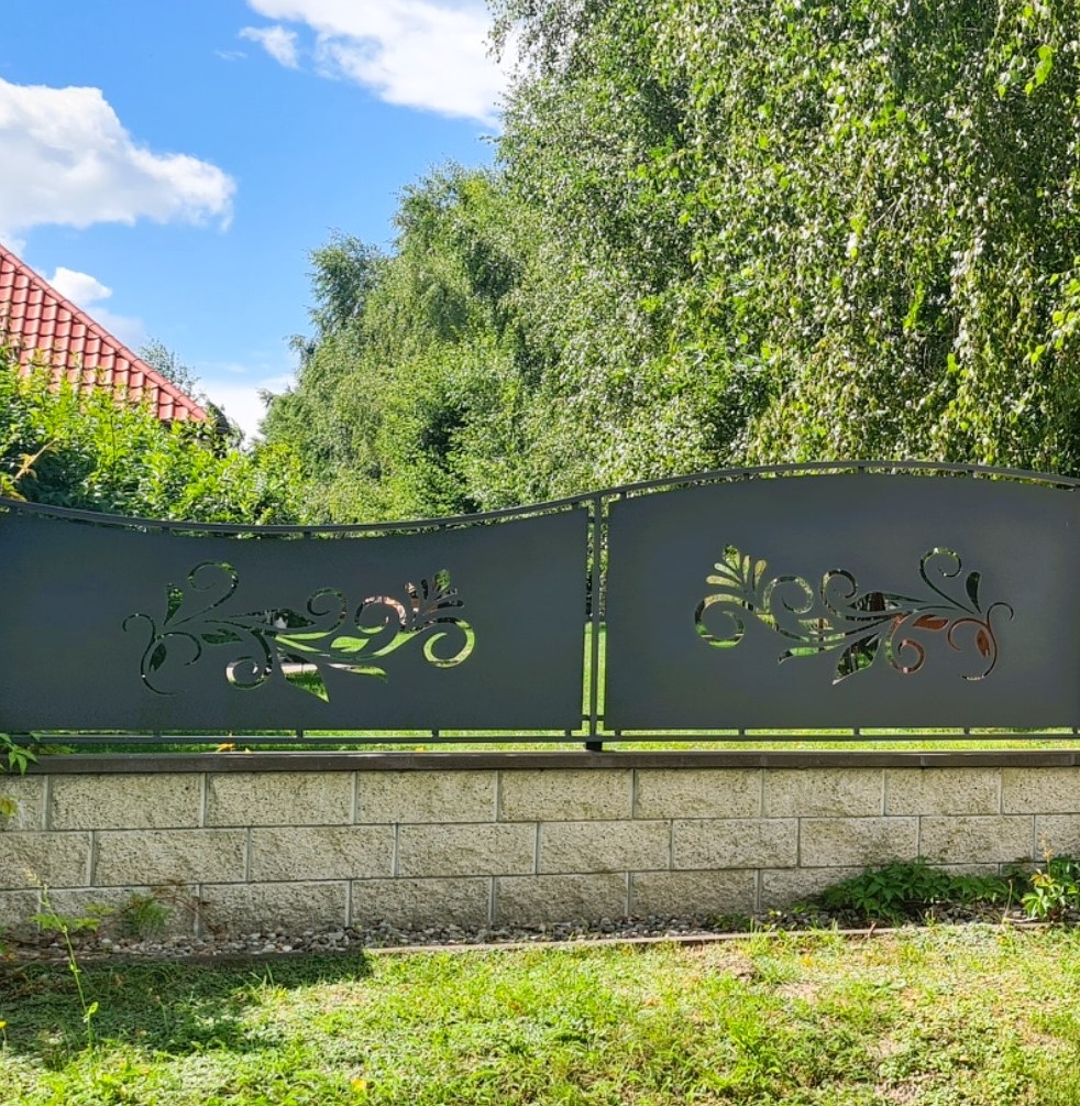 Czarny metalowy płot z ozdobnymi wycinanymi wzorami roślinnymi, zamontowany na murku z kamienia łupanego, na tle zieleni i fragmentu dachu.