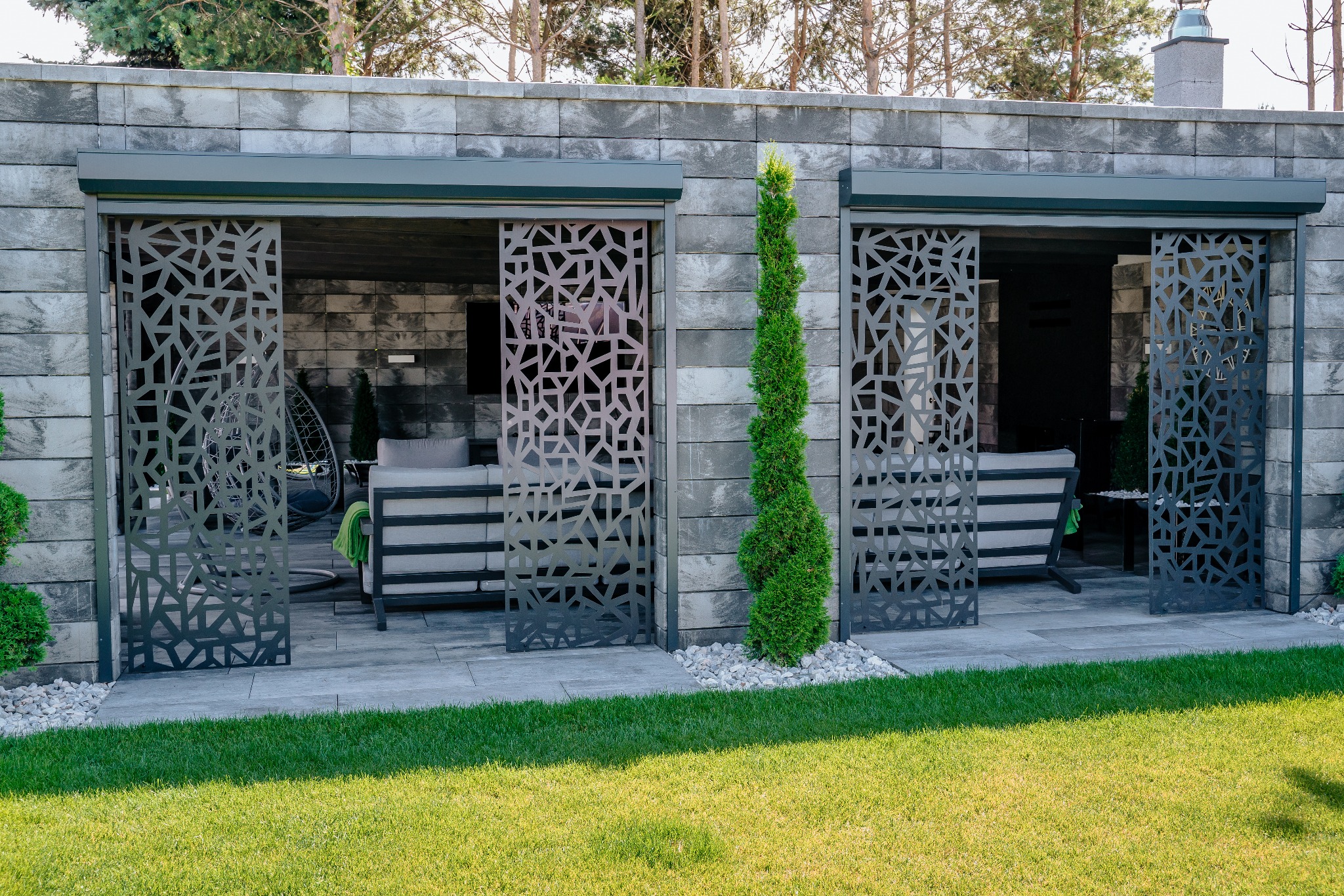 Nowoczesna altana z przesuwanymi, ażurowymi panelami metalowymi, z meblami ogrodowymi w środku, otoczona trawnikiem i zielenią.