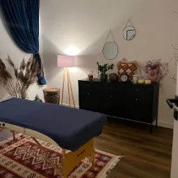 Pokój do zabiegów z łóżkiem do masażu przykrytym granatowym materiałem, wzorzystym dywanem, czarną komodą z dekoracjami i zasłoniętym oknem.