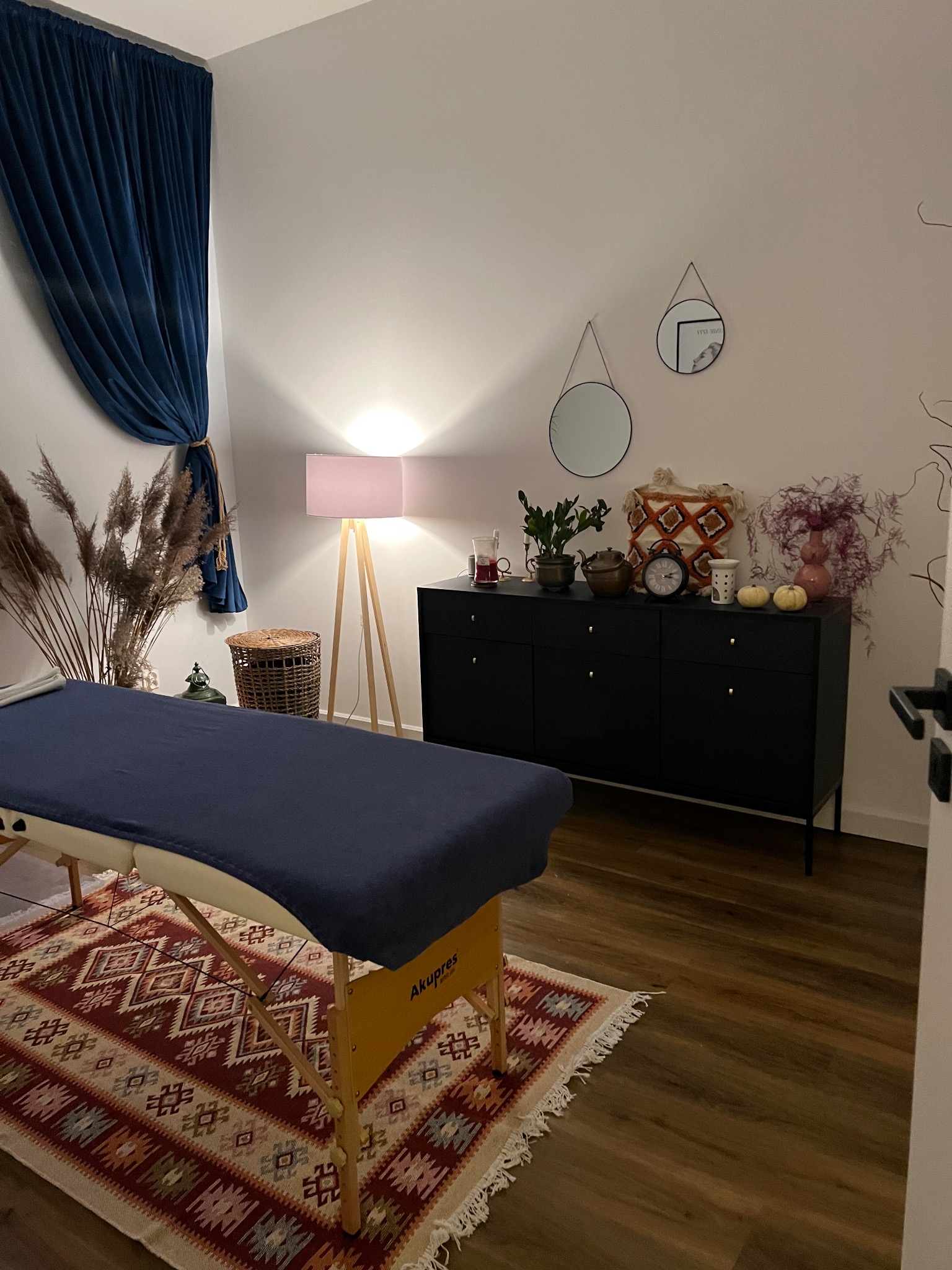 Pokój do zabiegów z łóżkiem do masażu przykrytym granatowym materiałem, wzorzystym dywanem, czarną komodą z dekoracjami i zasłoniętym oknem.