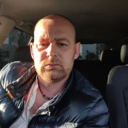 Portret mężczyzny z brodą w samochodzie, ubrany w błyszczącą, granatową kurtkę puchową i białą koszulę, oświetlony popołudniowym słońcem.