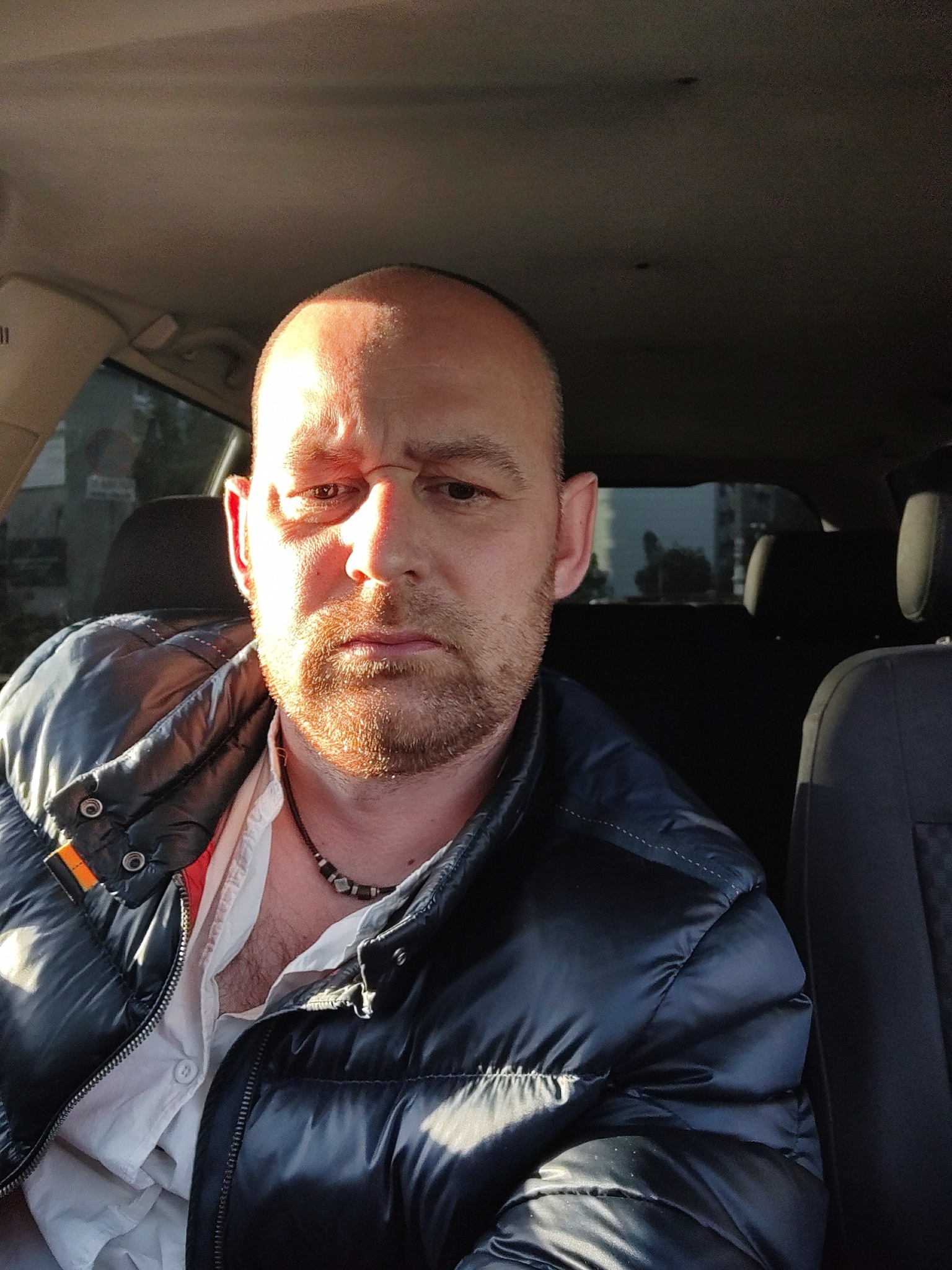 Portret mężczyzny z brodą w samochodzie, ubrany w błyszczącą, granatową kurtkę puchową i białą koszulę, oświetlony popołudniowym słońcem.