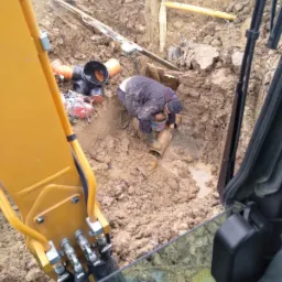 Pracownik w wykopie łączy rury kanalizacyjne, widok z kabiny koparki, błotniste otoczenie.