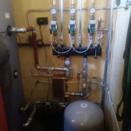 Rozdzielacz ogrzewania podłogowego z pompami obiegowymi, manometrami i rurami, zamontowany na żółtej ścianie w kotłowni.