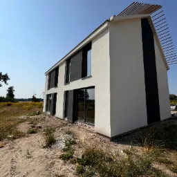 Nowoczesny dom z białą elewacją i czarnymi akcentami, widoczny z zewnątrz, z elementami drewnianymi na dachu, na tle błękitnego nieba i dzikiej roślinności.