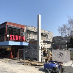 C77 sp.z.o.o - Budynek w trakcie budowy z czerwonym neonem 'SURFIN' na sąsiednim budynku, widoczne rusztowania i materiały budowlane, betonowy słup w centrum kadru.