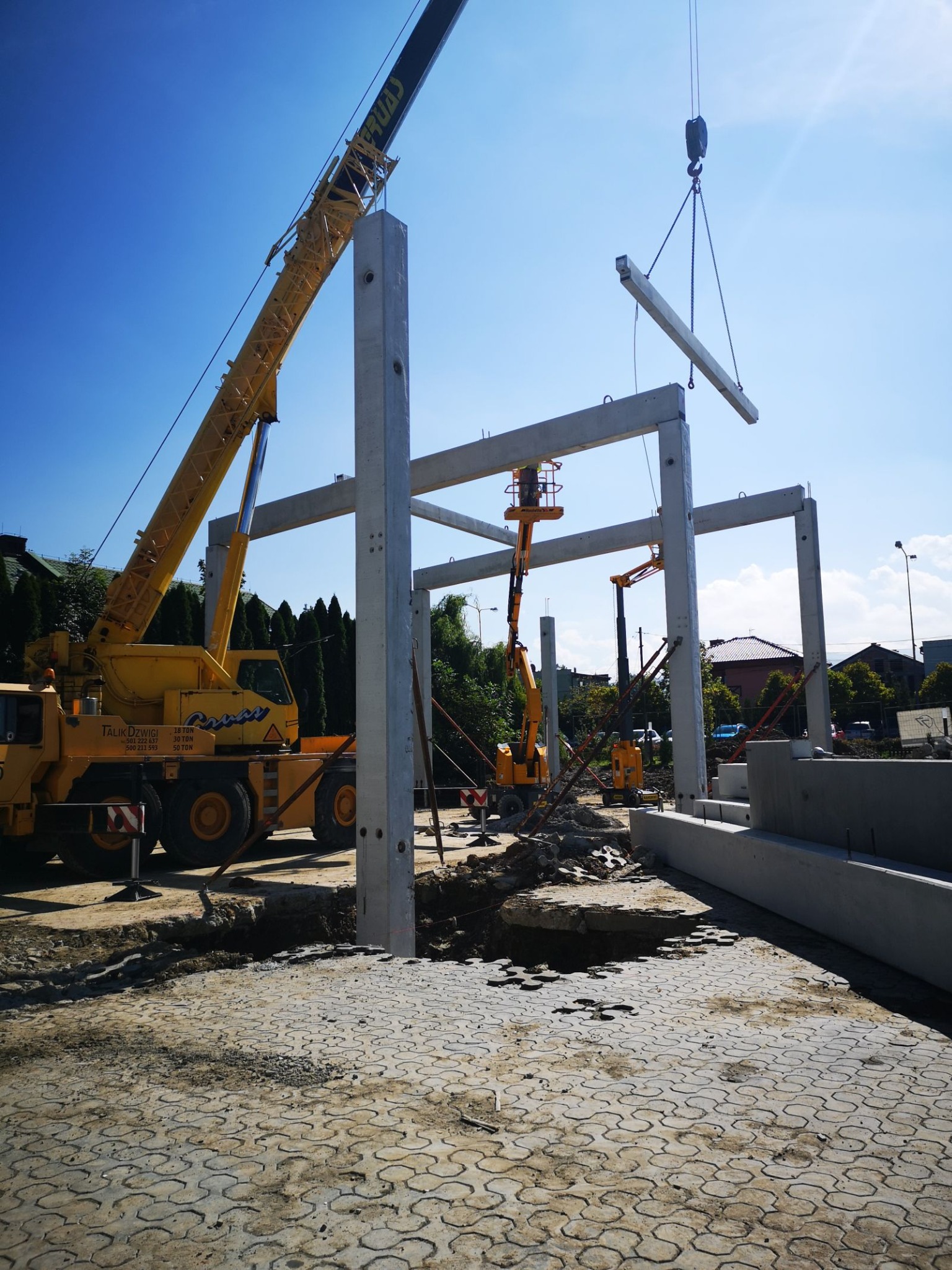 Montaż szkieletu domu z prefabrykatów betonowych przy użyciu dźwigu, widoczne słupy i belki, teren budowy z wykopami.