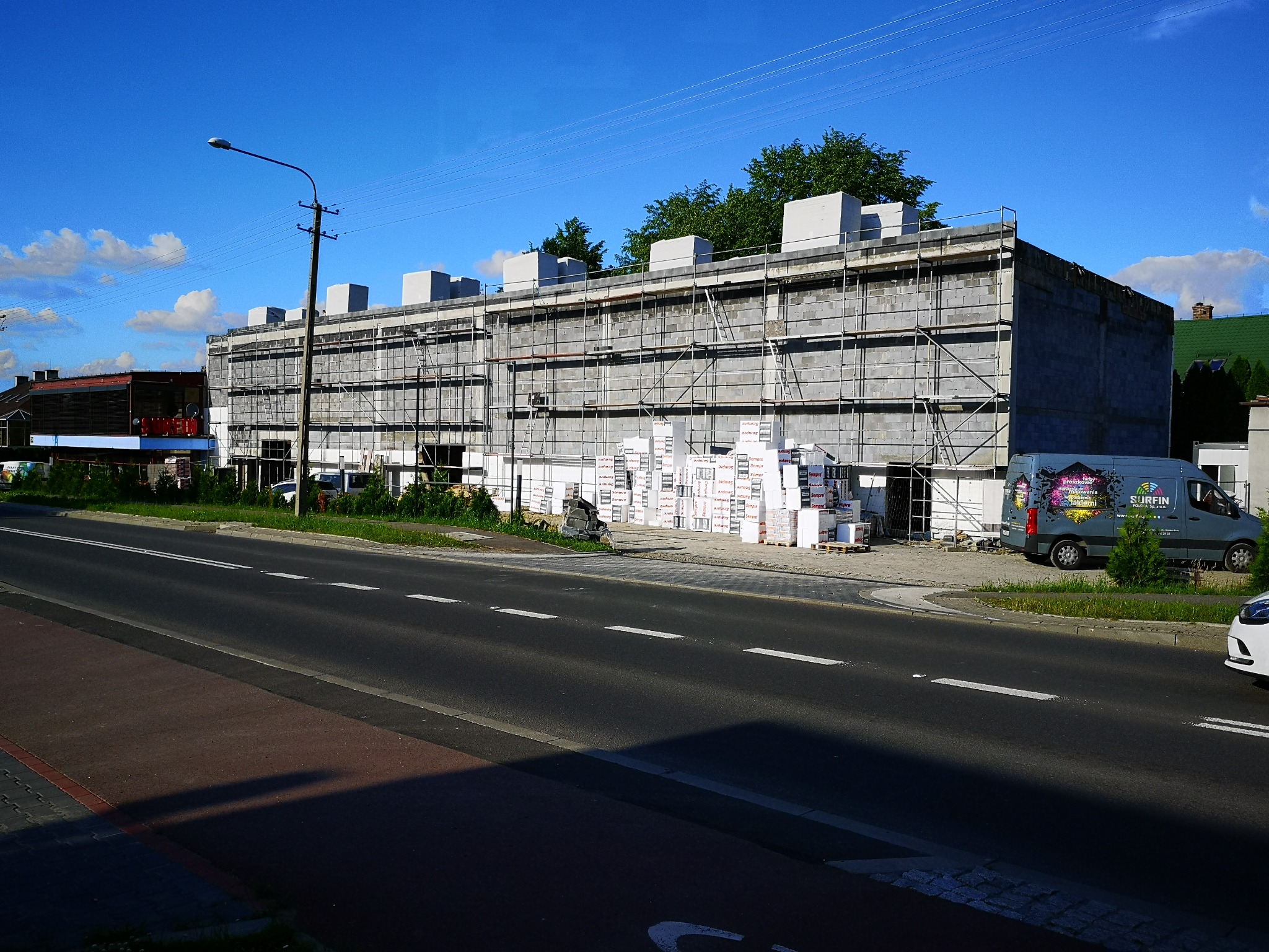 Budynek w trakcie budowy z szarych bloczków, otoczony rusztowaniami, z materiałami budowlanymi składowanymi obok i zaparkowanym samochodem dostawczym z reklamą firmy. Widok z ulicy w słoneczny dzień.