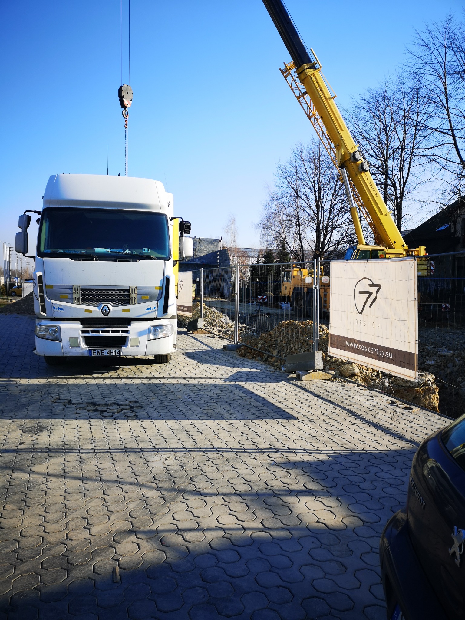 Ciężarówka Renault z dźwigiem budowlanym na placu budowy, widoczny banner z logo firmy projektowej Concept77, w tle wykop pod fundamenty ogrodzony siatką.