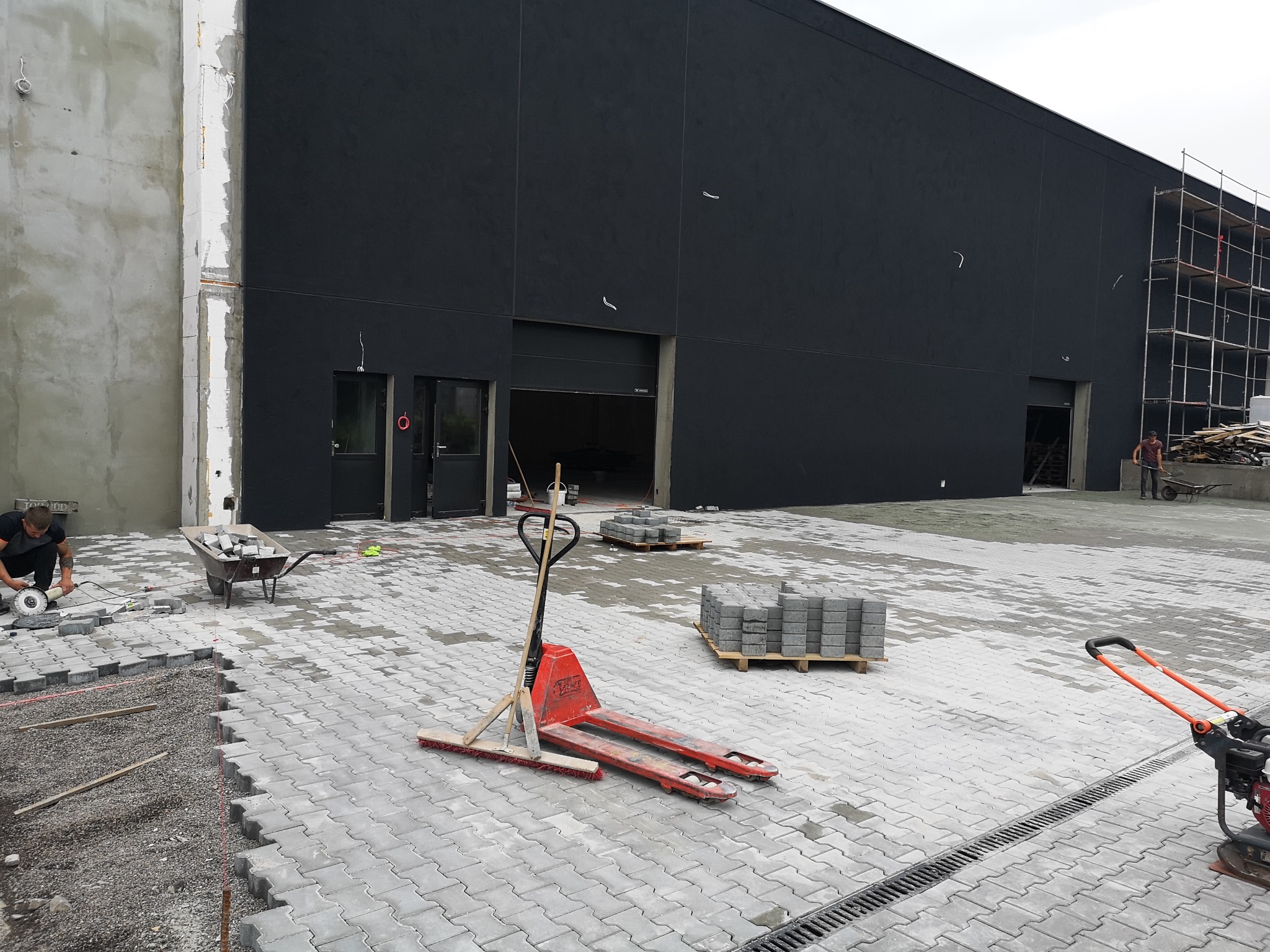 Układanie szarej kostki brukowej na posesji, widoczny pracownik przycinający kostkę szlifierką kątową, paleta z kostką, wózek widłowy ręczny i zagęszczarka wibracyjna.