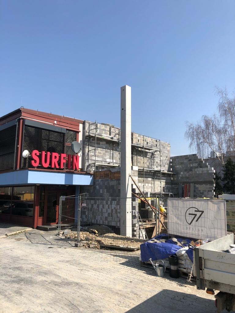 Budynek w trakcie budowy z czerwonym neonem 'SURFIN' na sąsiednim budynku, widoczne rusztowania i materiały budowlane, betonowy słup w centrum kadru.