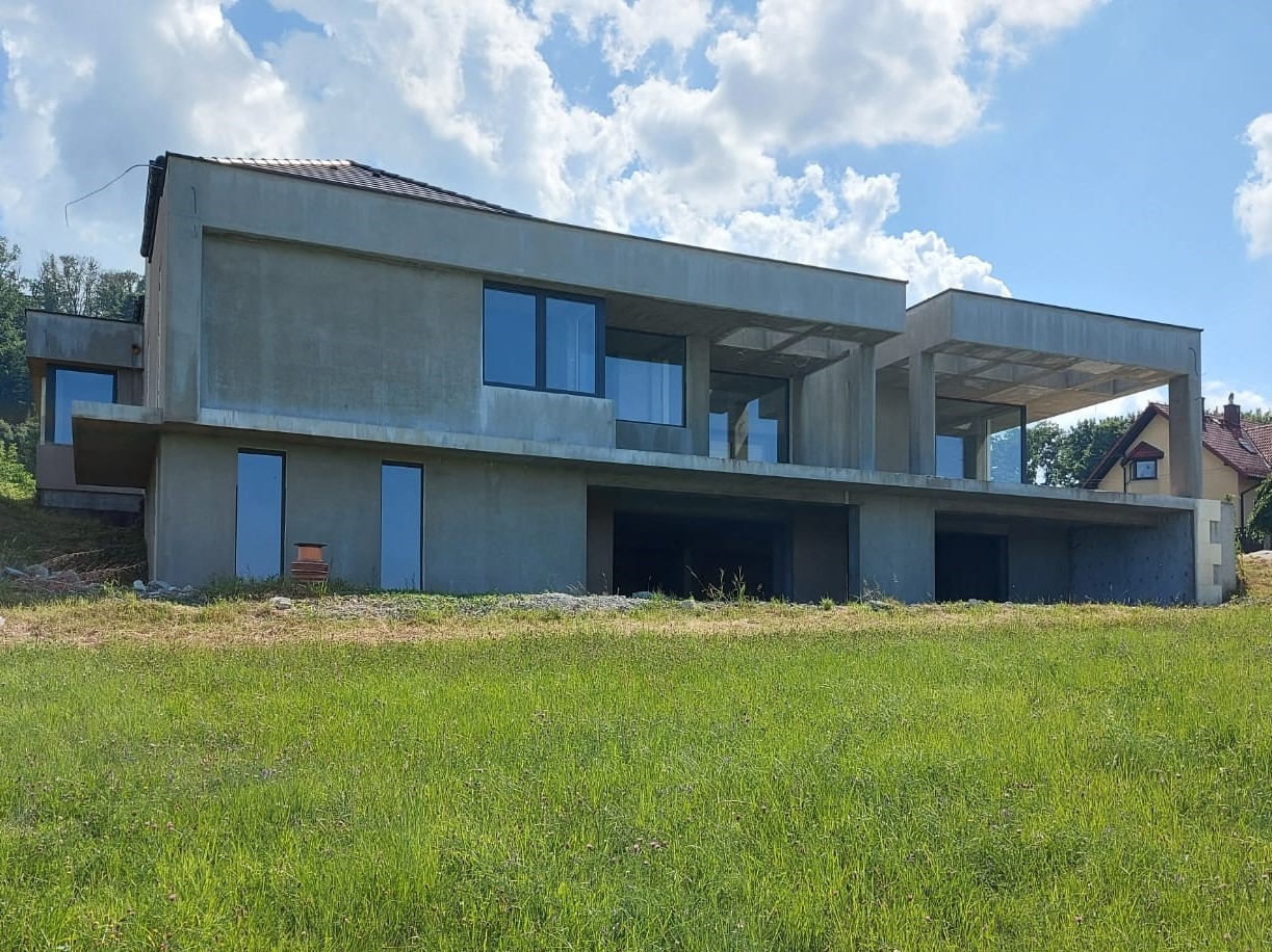 Surowy, dwukondygnacyjny dom murowany w stanie deweloperskim, z widocznymi otworami okiennymi i garażowymi, na tle zielonej łąki i błękitnego nieba z chmurami.