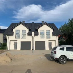 Nowoczesny bliźniak z szarą dachówką i garażami, widoczny z zewnątrz, z logo firmy budowlanej na zaparkowanym samochodzie.