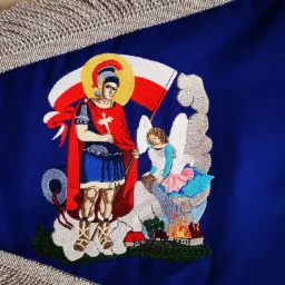 Dekoracyjny, trójkątny proporzec w kolorze granatowym, obramowany srebrnym frędzlem. Na tkaninie wyhaftowana postać rycerza z aureolą, trzymającego krzyż, oraz anioła nad płonącymi domami. Haft...