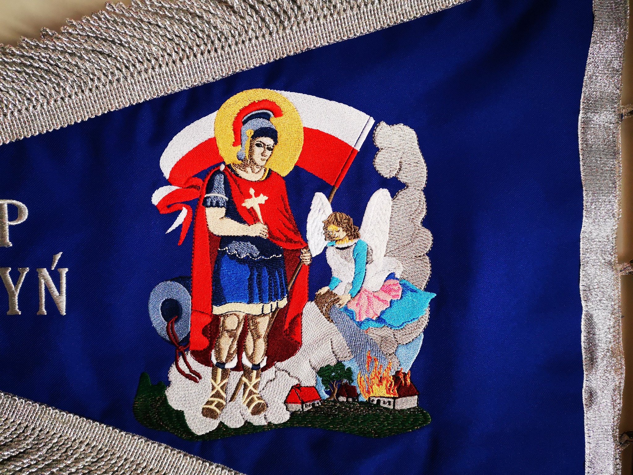 Dekoracyjny, trójkątny proporzec w kolorze granatowym, obramowany srebrnym frędzlem. Na tkaninie wyhaftowana postać rycerza z aureolą, trzymającego krzyż, oraz anioła nad płonącymi domami. Haft...