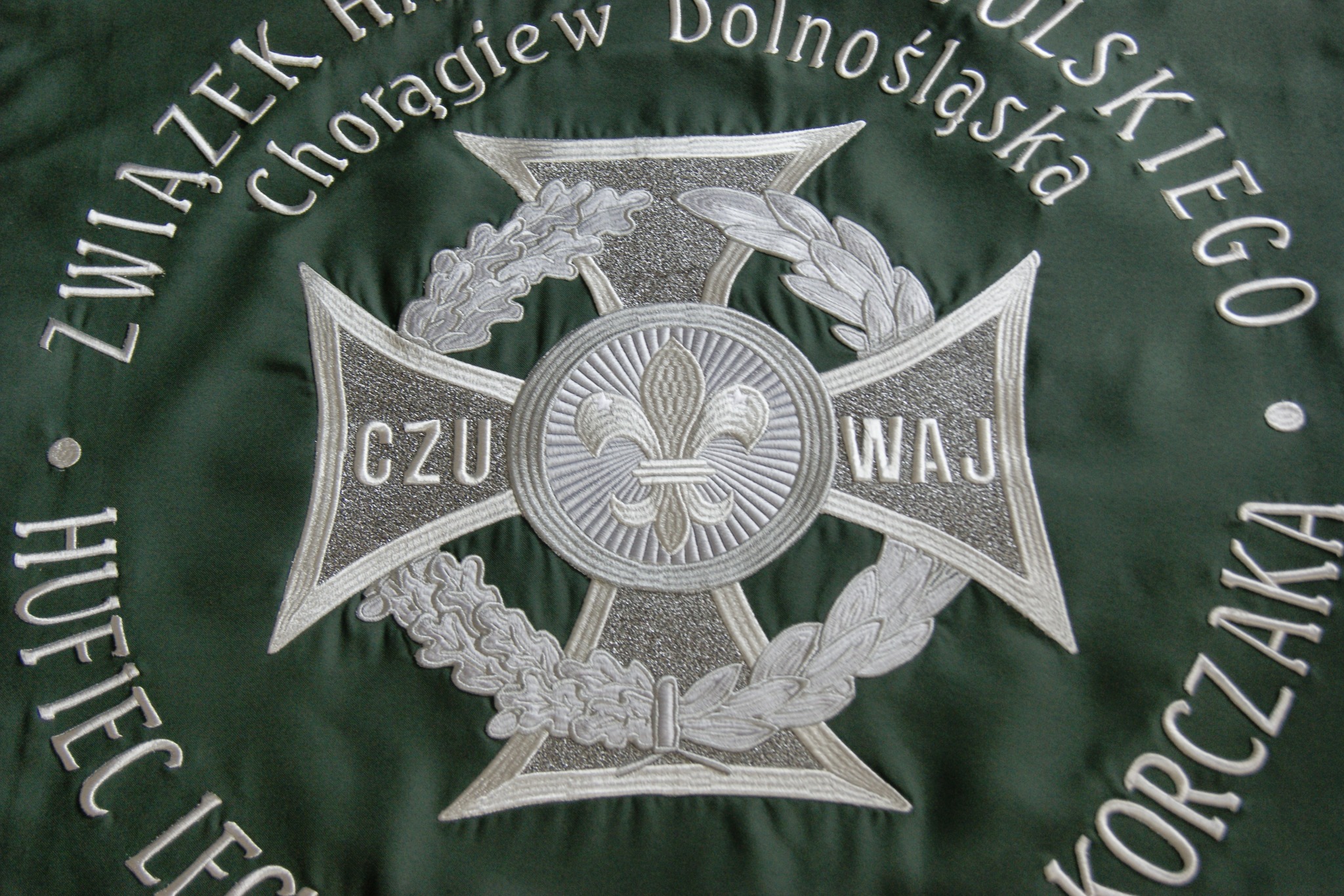 Zbliżenie na zielony sztandar harcerski z wyhaftowanym srebrnym krzyżem z lilijką i napisem 'Związek Harcerstwa Polskiego Chorągiew Dolnośląska Hufiec Legnica Korczak'