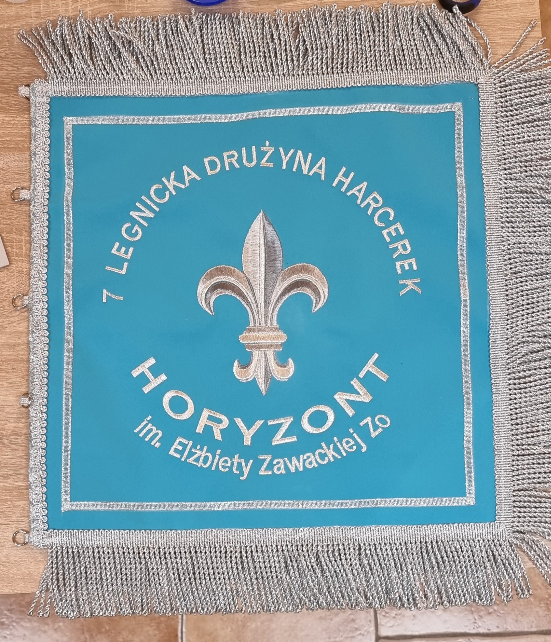 Błękitny proporzec z haftowanym srebrną nicią tekstem '7 Legnicka Drużyna Harcerek HORIZONT im. Elżbiety Zawackiej Zo' i symbolem lilijki, obramowany srebrnymi frędzlami.