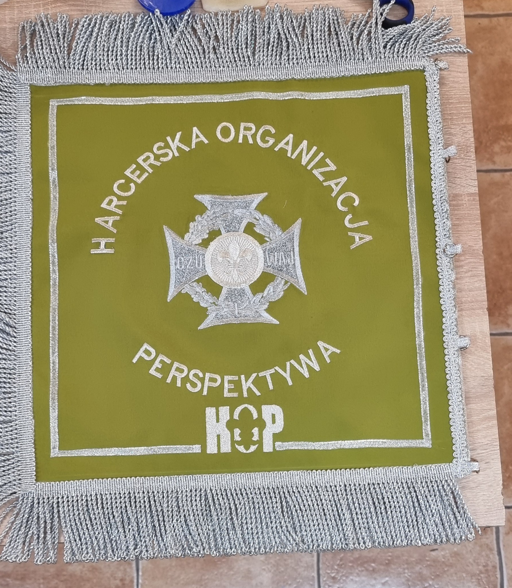 Z bliska widoczny jest sztandar harcerski w kolorze oliwkowym z srebrnym haftem, obramowany srebrnymi frędzlami.