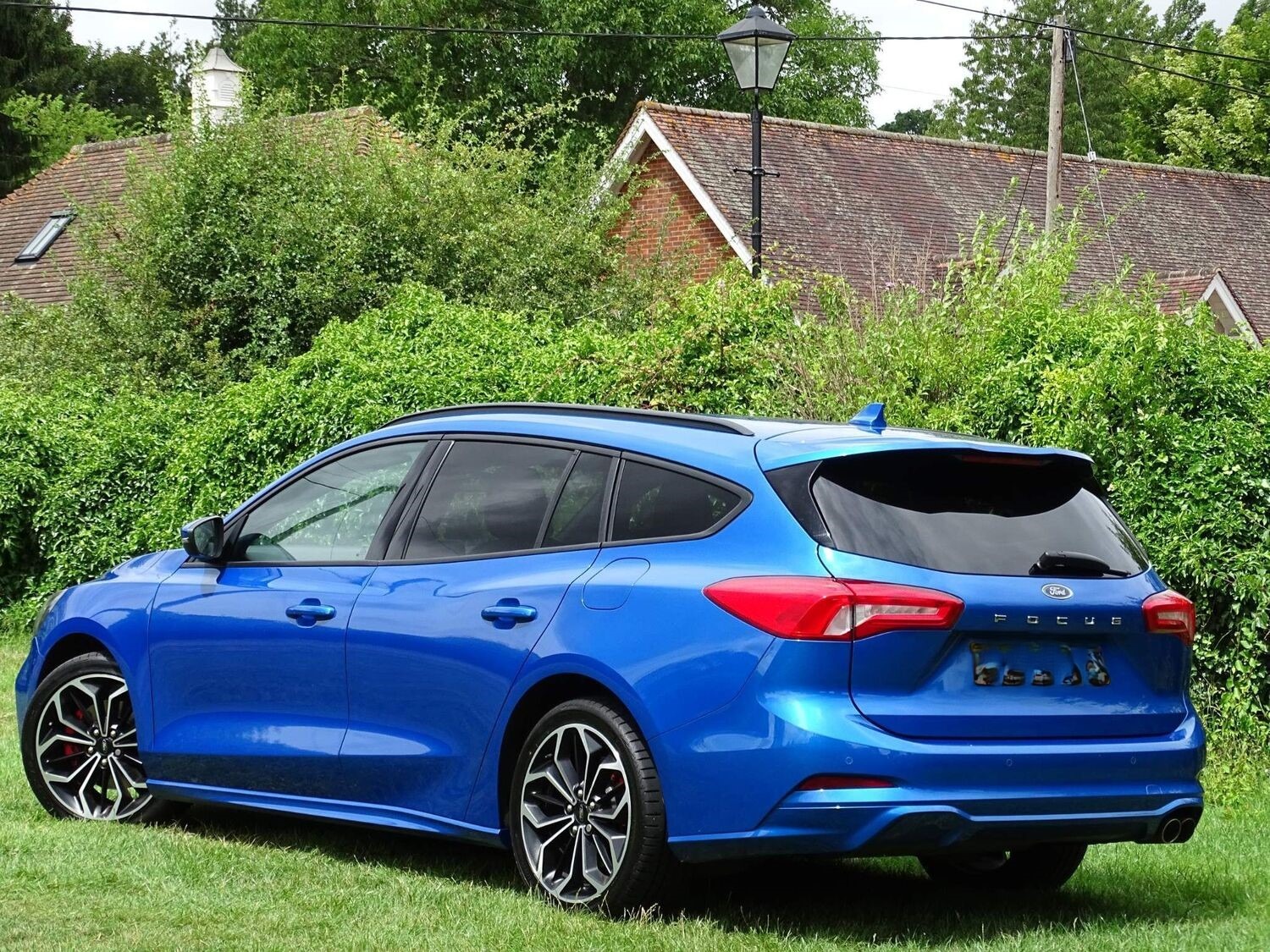 Niebieski Ford Focus kombi z czarnymi felgami, zaparkowany na trawniku przed budynkiem z czerwonej cegły i latarnią.