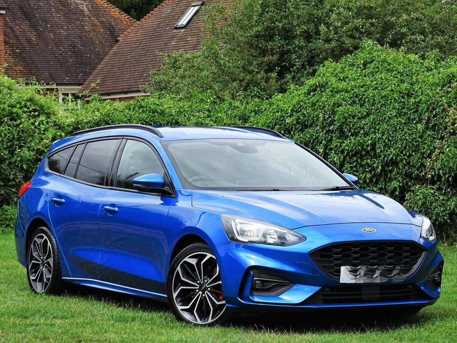 Niebieski samochód kombi marki Ford Focus ST na trawniku przed domem z czerwonym dachem i zielonym żywopłotem w tle.