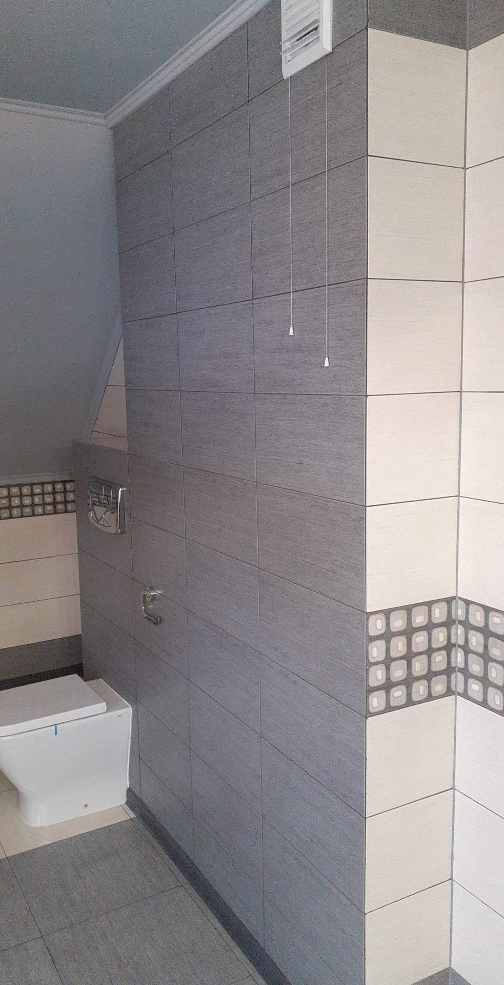 Wykończona toaleta w łazience na poddaszu, z szarymi i kremowymi płytkami, dekoracyjnym pasem z motywem geometrycznym oraz widoczną wentylacją sufitową.