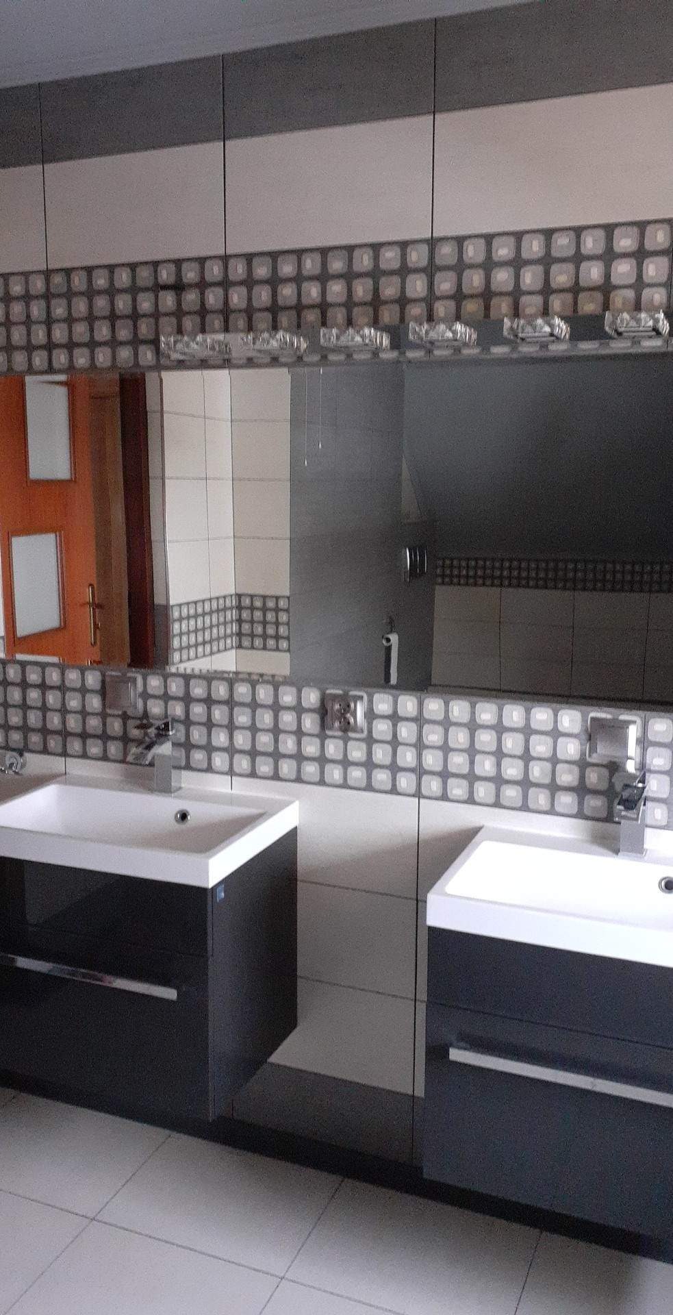 Umywalki z szafkami w ciemnym kolorze w łazience wykończonej płytkami w odcieniach szarości i bieli, nad umywalkami lustro z dekoracyjnym oświetleniem.