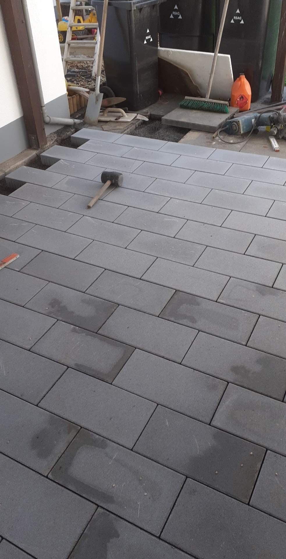 Układanie szarej kostki brukowej na tarasie, widoczny gumowy młotek, fragmenty niezakończonej powierzchni, narzędzia i pojemniki na śmieci w tle.