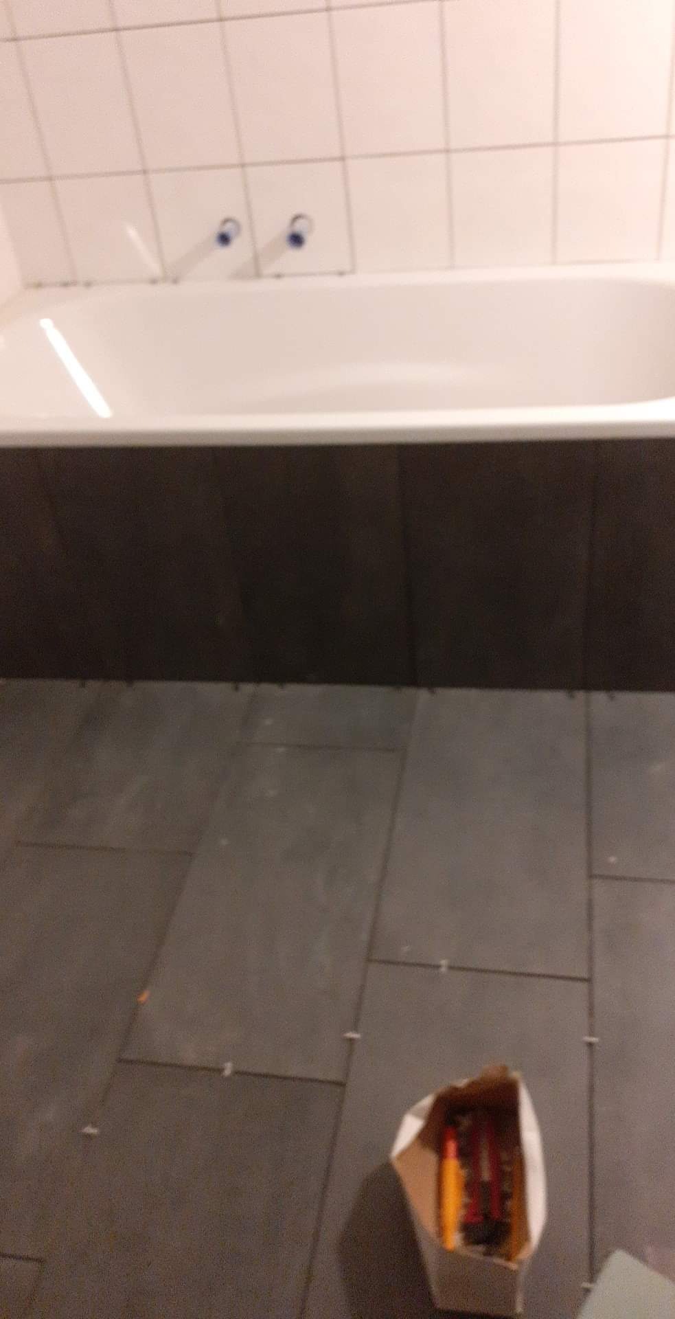 Fragment łazienki w trakcie układania ciemnych płytek podłogowych, widoczna wanna z białymi płytkami na ścianie i pudełko z narzędziami.