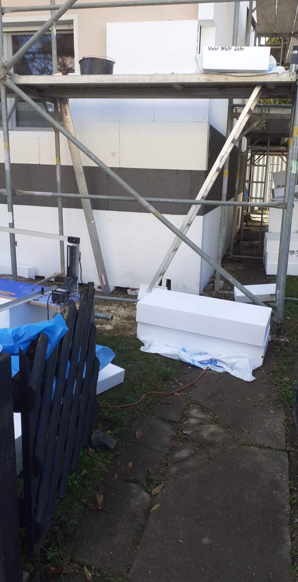 Ocieplanie elewacji budynku styropianem, widoczne rusztowanie, płyty styropianowe oraz narzędzia na zewnątrz budynku.