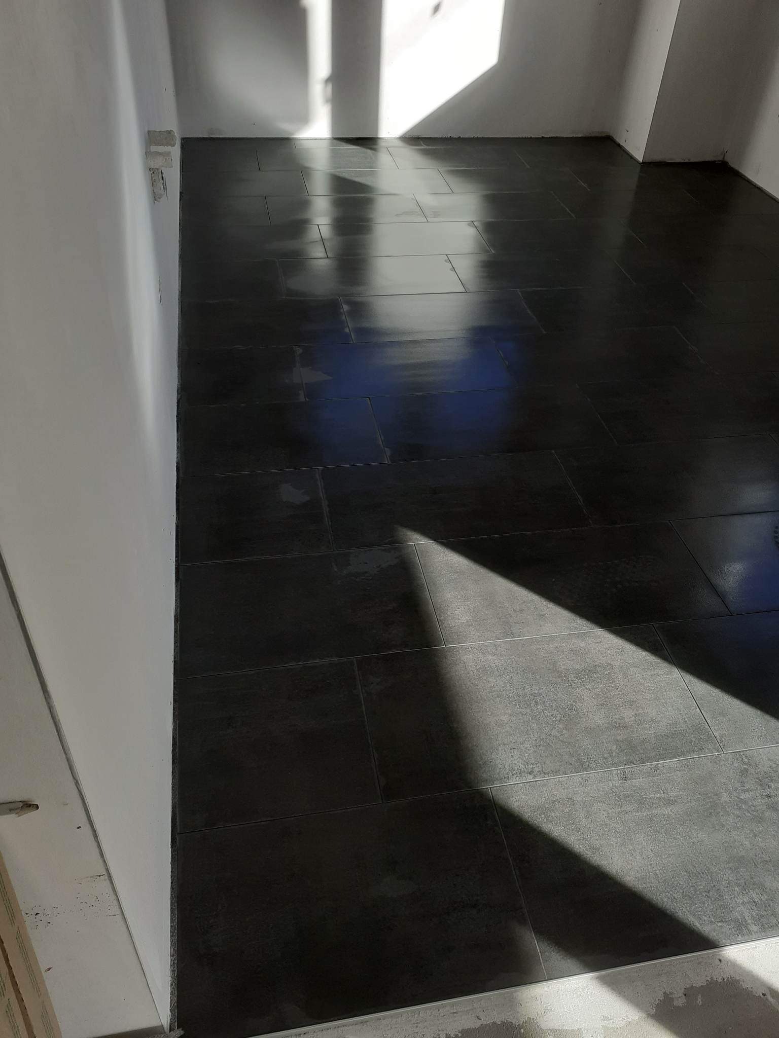 Ułożone, ciemne płytki podłogowe w pomieszczeniu w trakcie prac wykończeniowych, widoczne niedoskonałości na ścianach i intensywne światło słoneczne padające na podłogę.