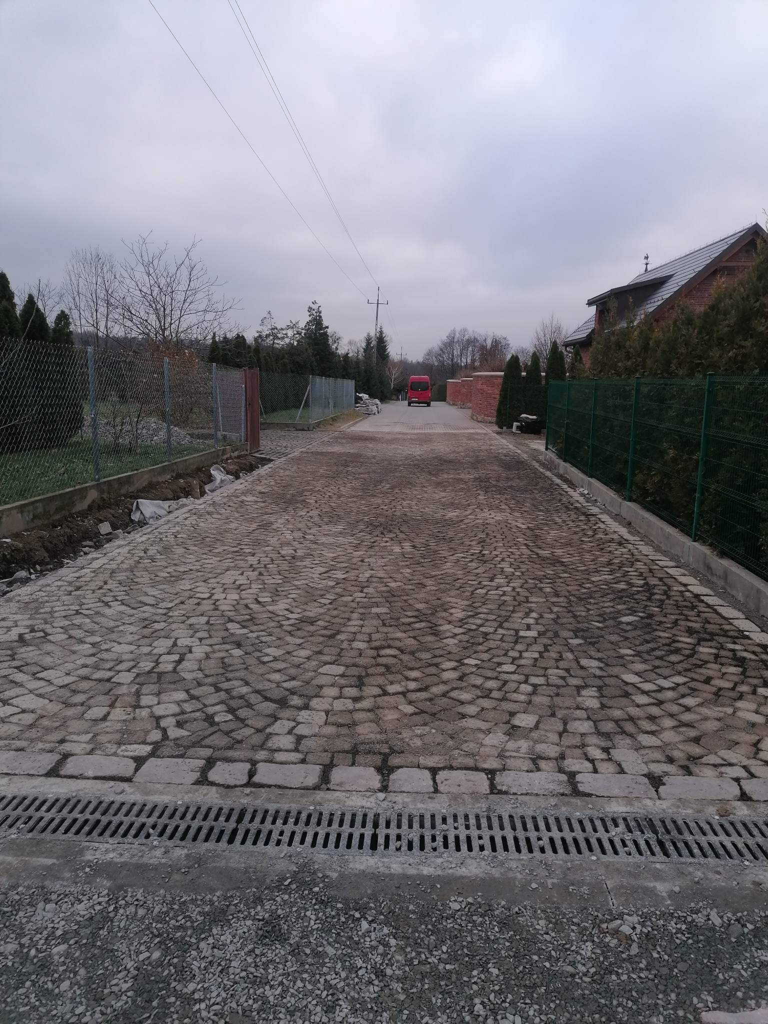 Ułożona nawierzchnia z kostki granitowej w kształcie wachlarza, widoczny odpływ liniowy i przygotowane podłoże pod obrzeża, w tle czerwony bus i dom z ciemną dachówką.