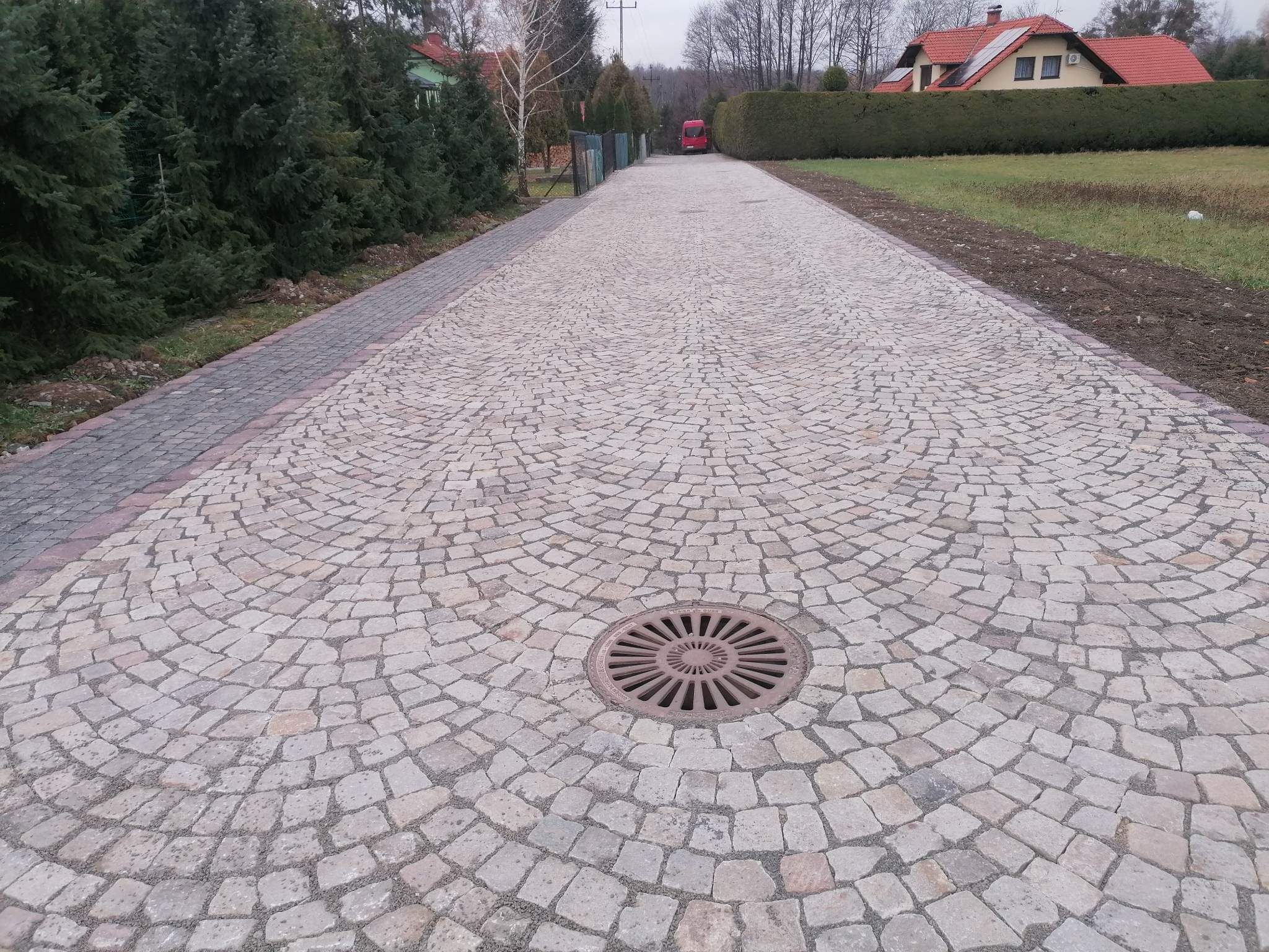 Długa aleja z jasnoszarej kostki granitowej ułożonej w wachlarzowy wzór, z centralnie umieszczoną studzienką kanalizacyjną, otoczona zielenią i domami w tle.