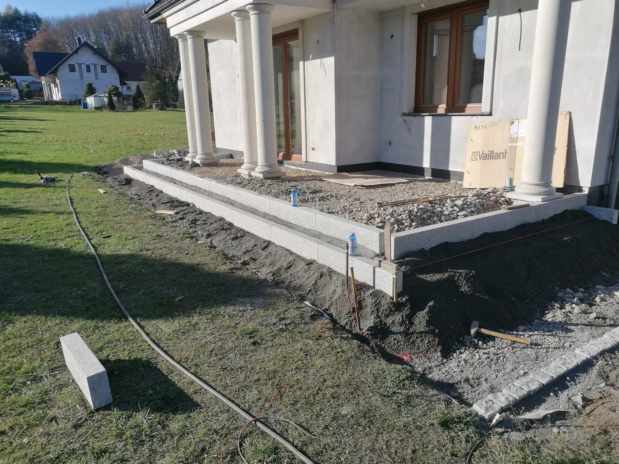 Budowa tarasu: dwupoziomowa konstrukcja z jasnoszarej kostki granitowej w trakcie montażu, widoczne narzędzia i materiały budowlane, trawnik i dom z kolumnami w tle.