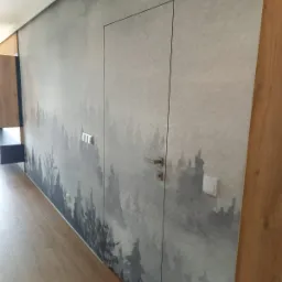 Ściana z fototapetą przedstawiającą motyw leśny, z ukrytymi drzwiami z klamką i włącznikami światła, po prawej stronie widoczna drewniana boazeria.