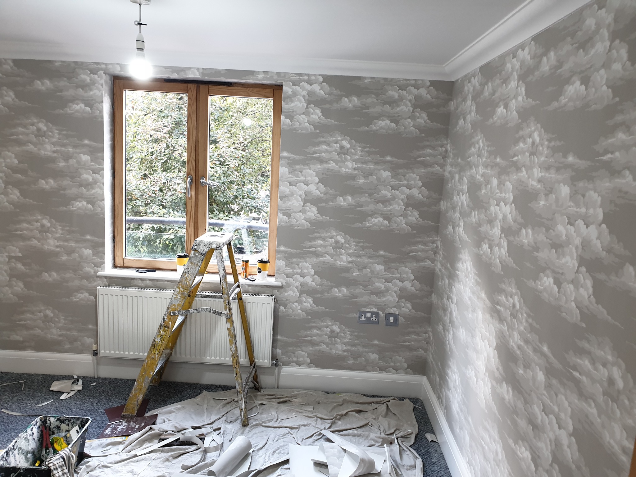 Pokój po tapetowaniu w motyw chmur, widoczna drabina, przykryte podłoże, tapeta szaro-biała.