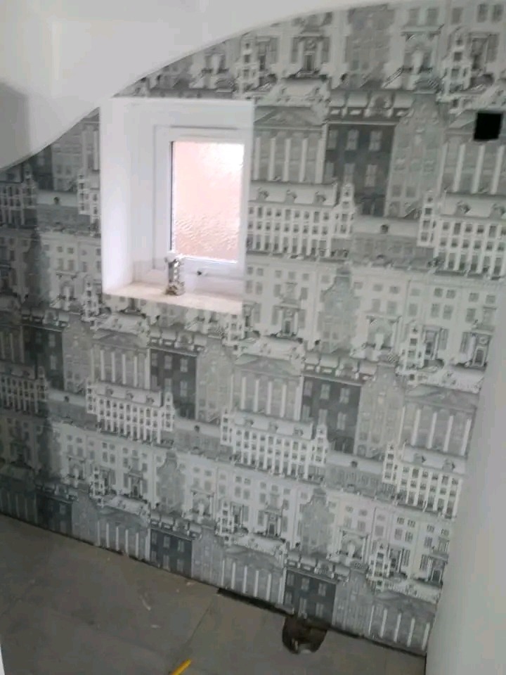 Ściana pokryta tapetą z motywem budynków w odcieniach szarości, obok okno z białym obramowaniem. Na podłodze widoczny fragment żółtego narzędzia.