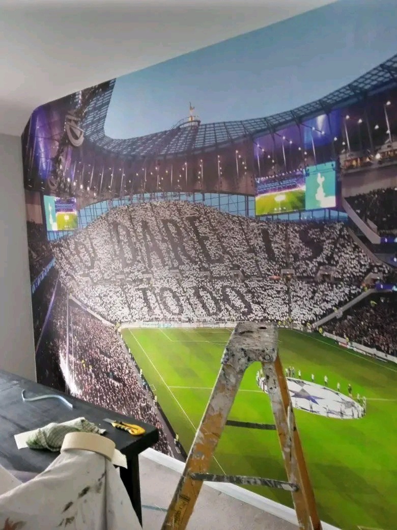 Fragment pokoju z nałożoną tapetą przedstawiającą stadion Tottenham Hotspur, widoczna drabina malarska i akcesoria do tapetowania na stole.