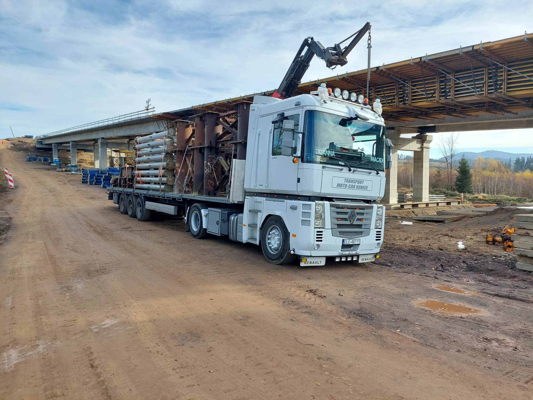 Biała ciężarówka Renault z dźwigiem przewozi elementy konstrukcyjne mostu na platformie, widok z boku na tle budowanego wiaduktu i jesiennego krajobrazu.