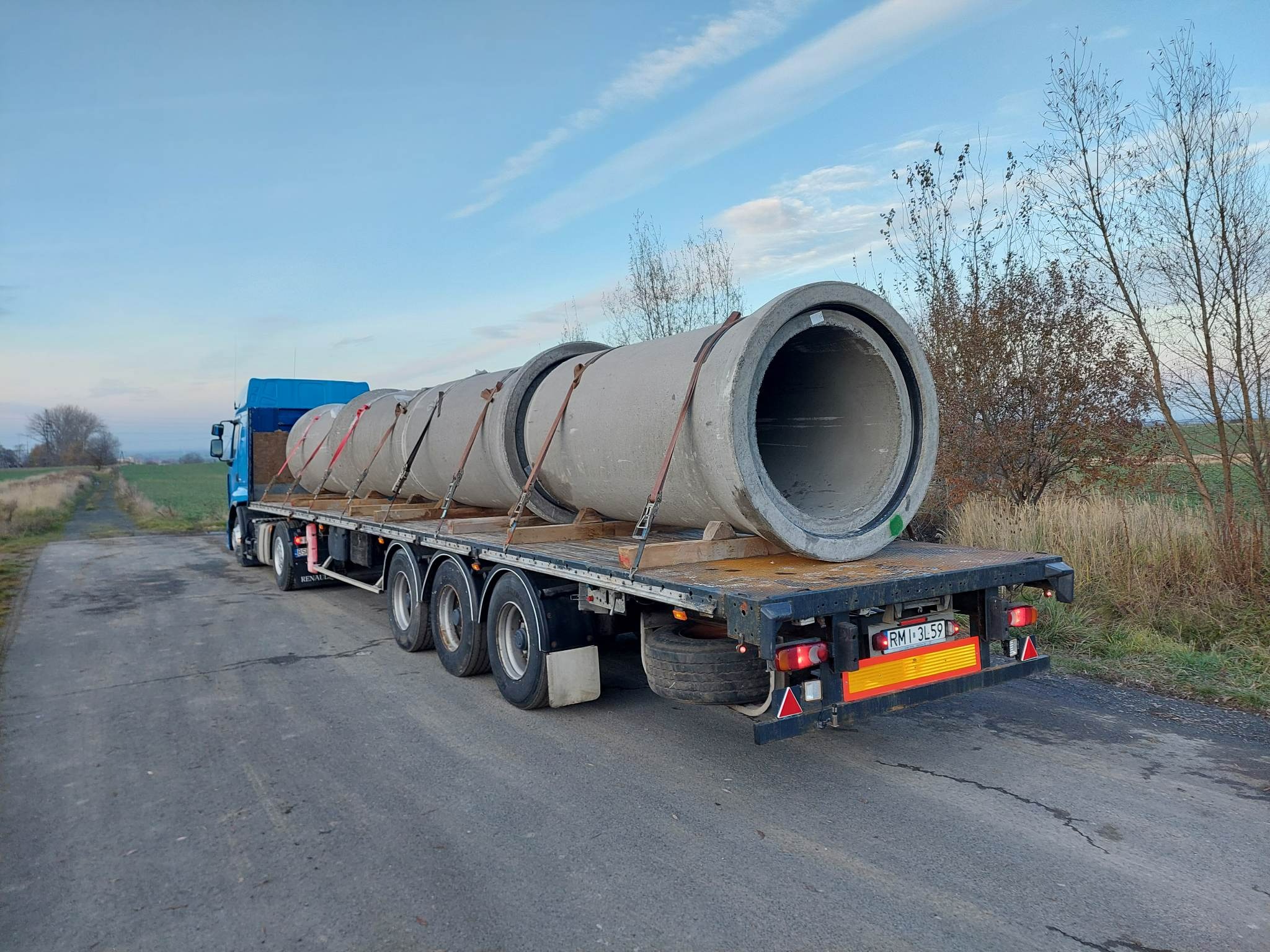 Niebieska ciężarówka z naczepą przewożąca cztery betonowe rury zabezpieczone pasami transportowymi na tle błękitnego nieba z chmurami i jesiennego krajobrazu.