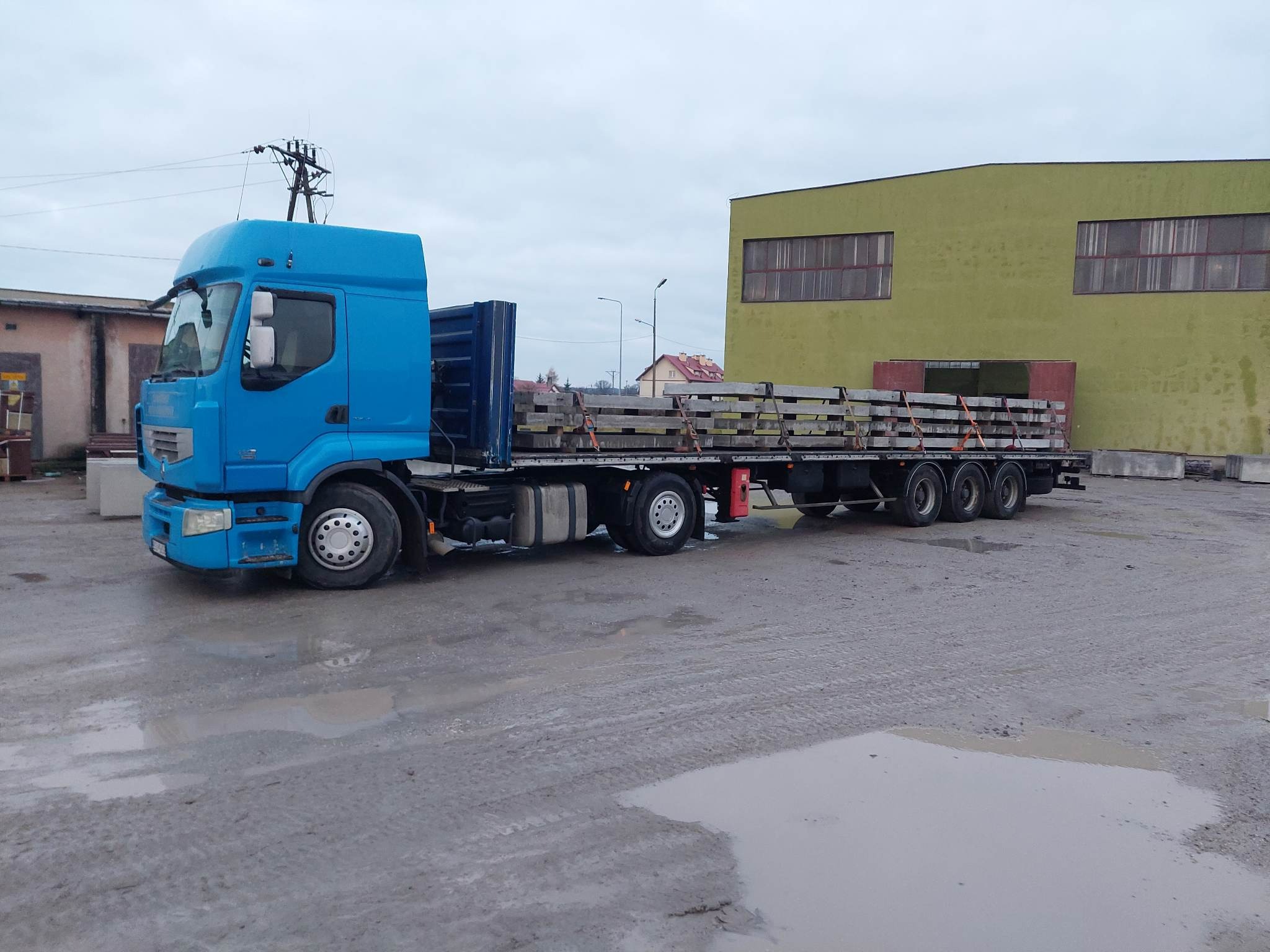 Niebieska ciężarówka z platformą załadowana prefabrykatami betonowymi, zaparkowana na placu budowy w pochmurny dzień.