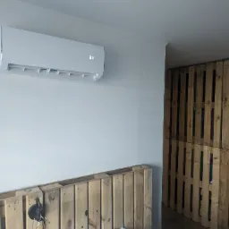 Biała klimatyzacja typu split zamontowana na ścianie w sypialni z zagł&oacute;wkiem z palet, wyświetlająca temperaturę 31 stopni Celsjusza, obok ściana z palet.