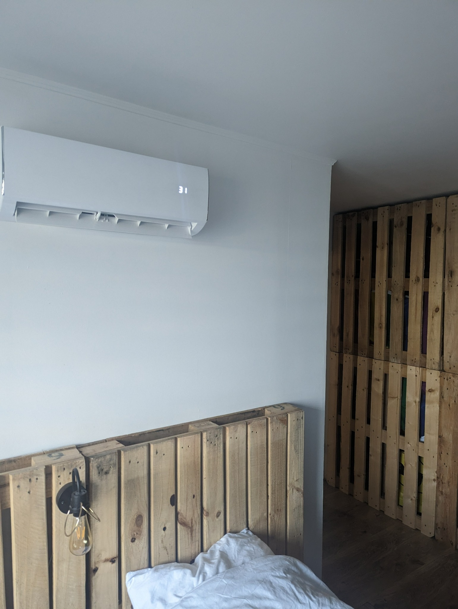 Biała klimatyzacja typu split zamontowana na ścianie w sypialni z zagłówkiem z palet, wyświetlająca temperaturę 31 stopni Celsjusza, obok ściana z palet.