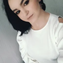 Portret kobiety z ciemnymi włosami, w białej bluzce z odkrytymi ramionami i złotych kolczykach w kształcie krzyżyków.