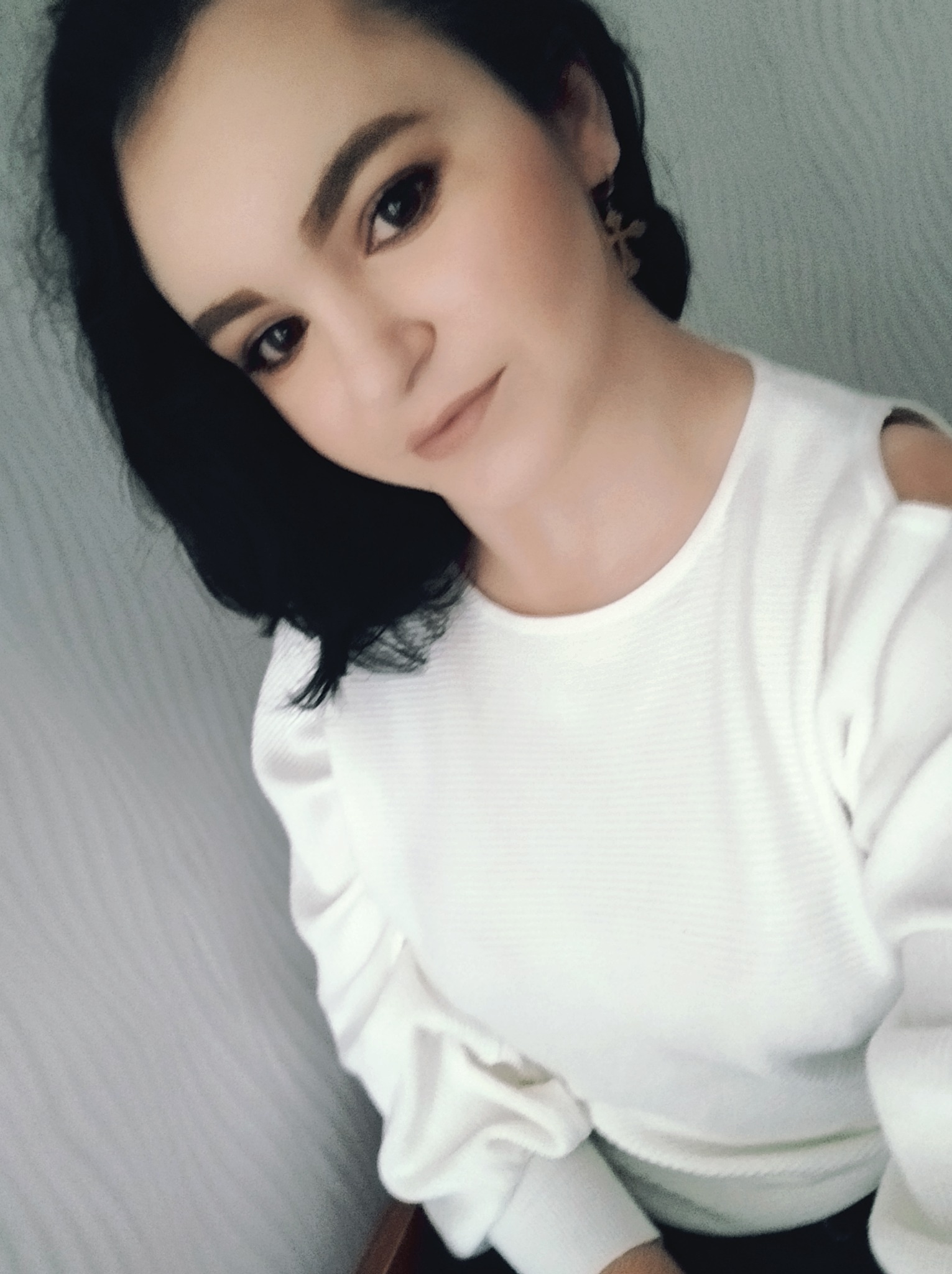 Portret kobiety z ciemnymi włosami, w białej bluzce z odkrytymi ramionami i złotych kolczykach w kształcie krzyżyków.