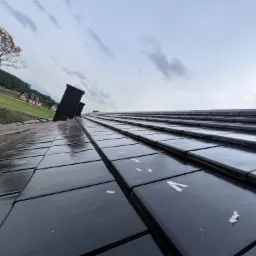 Nowy dach z czarnej dachówki ceramicznej, widok pod kątem, z widocznym krajobrazem w tle i fragmentami uszkodzeń na powierzchni dachówek.