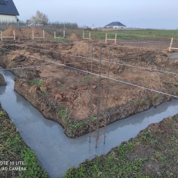 Usługi Ogólnobudowlane Maciej Bąk - Wylane fundamenty w wykopie, zbrojenie stalowe w narożniku, widoczne sznurki wyznaczające granice, w tle ziemia i domy.