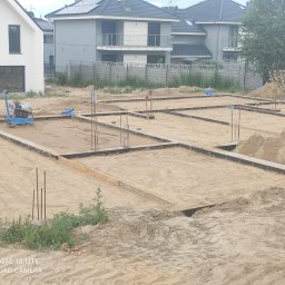 Usługi Ogólnobudowlane Maciej Bąk - Przygotowanie terenu pod budowę: wykopane fundamenty z szalunkiem, wystające pręty zbrojeniowe, ubijarka wibracyjna w tle, nowe domy jednorodzinne w tle.
